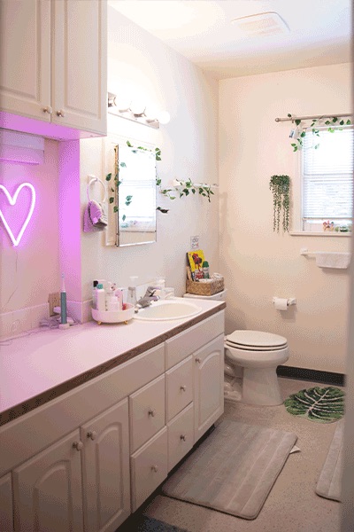 A bright bathroom with abundant white cabinets, a neon heart light, toilet, sink, mirror and green foliage decor.
